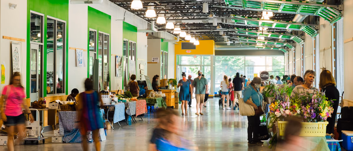 The indoor Farm Fresh Rhode Island market hall. Shoppers move between trestle tables of produce under a glazed roof and green-painted steel, a florist’s buckets of cut flowers in the foreground and a coffee stand at the far end.