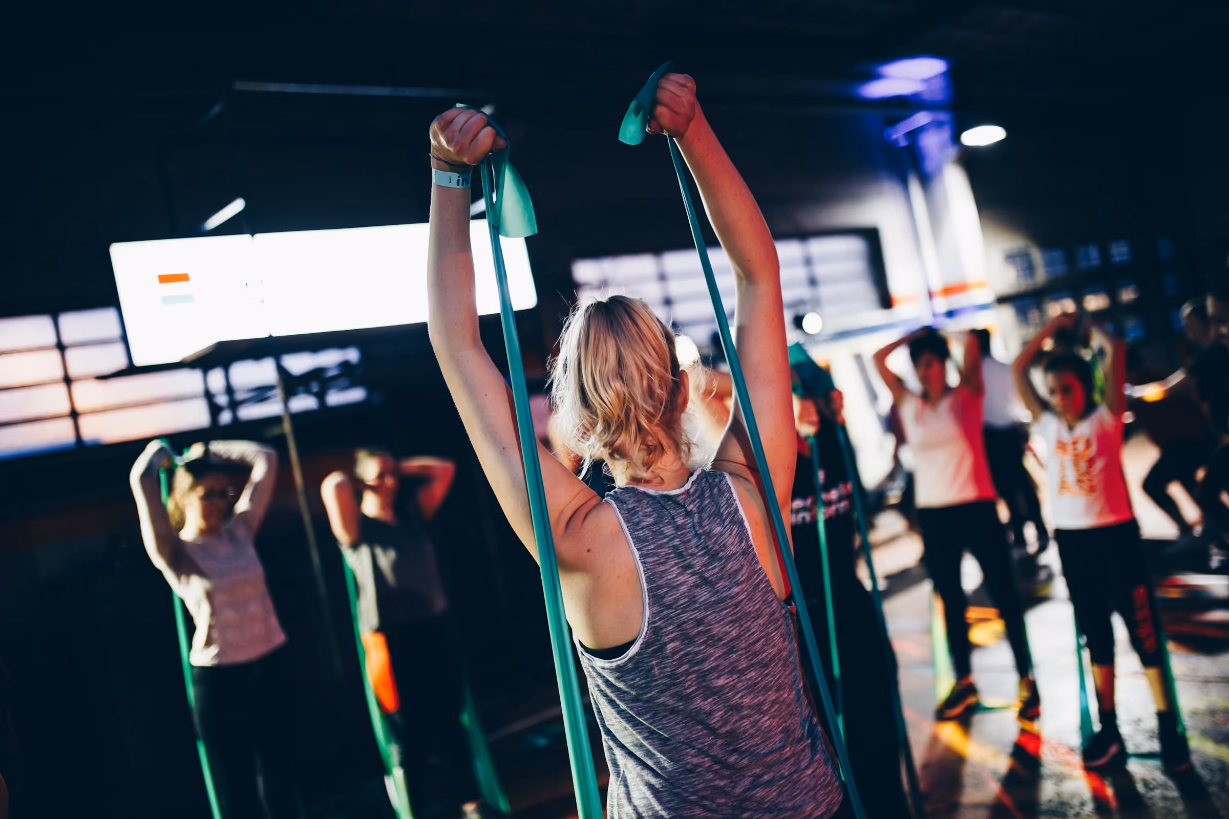 A group fitness class mid-workout, seen from within the crowd: people stretching resistance bands overhead in a large, warmly lit gym.
