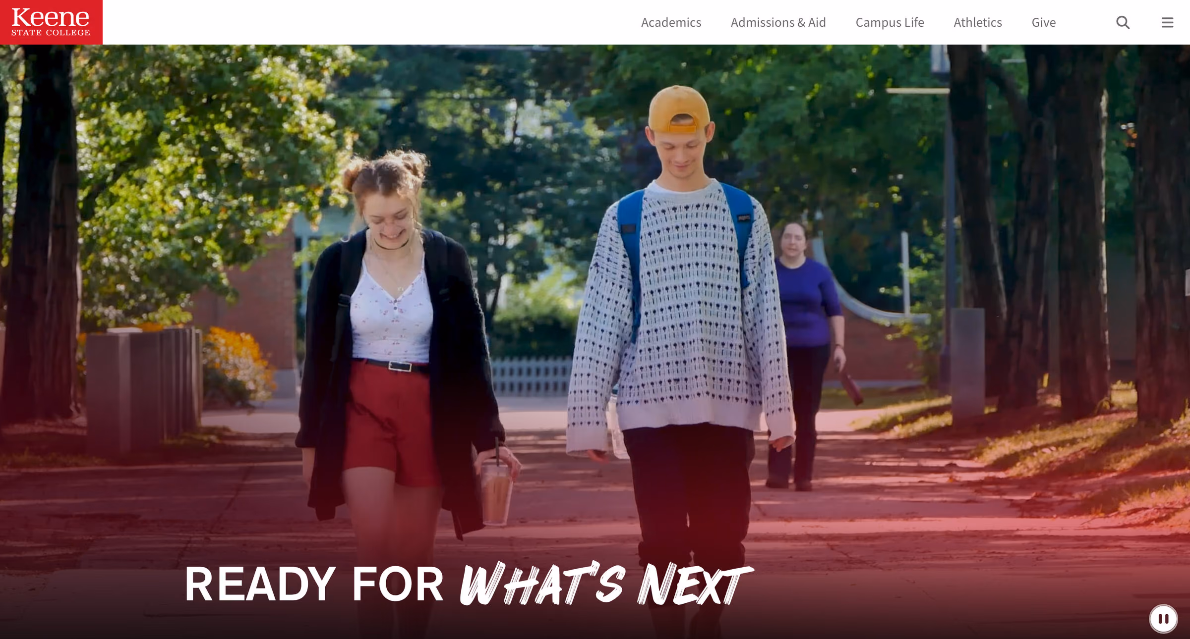 The Keene State College homepage. A five-item main navigation reads Academics, Admissions & Aid, Campus Life, Athletics, Give, beside a search control. Below it a full-width campus video shows two students walking a tree-lined path, captioned “Ready for What’s Next”.