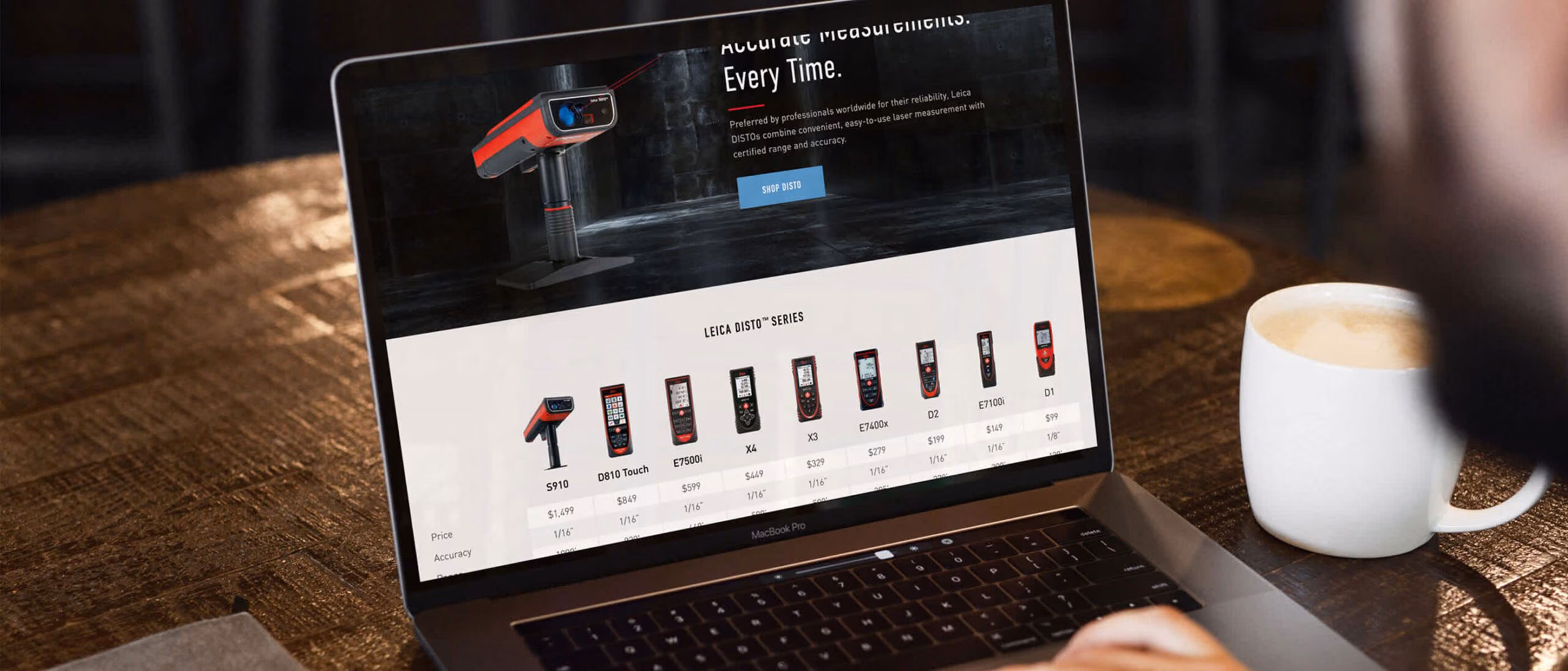 Over a shoulder at a wooden cafe table: a laptop showing the Leica DISTO shop — “Accurate Measurements. Every Time.” over a product comparison row with prices — beside a cup of coffee.