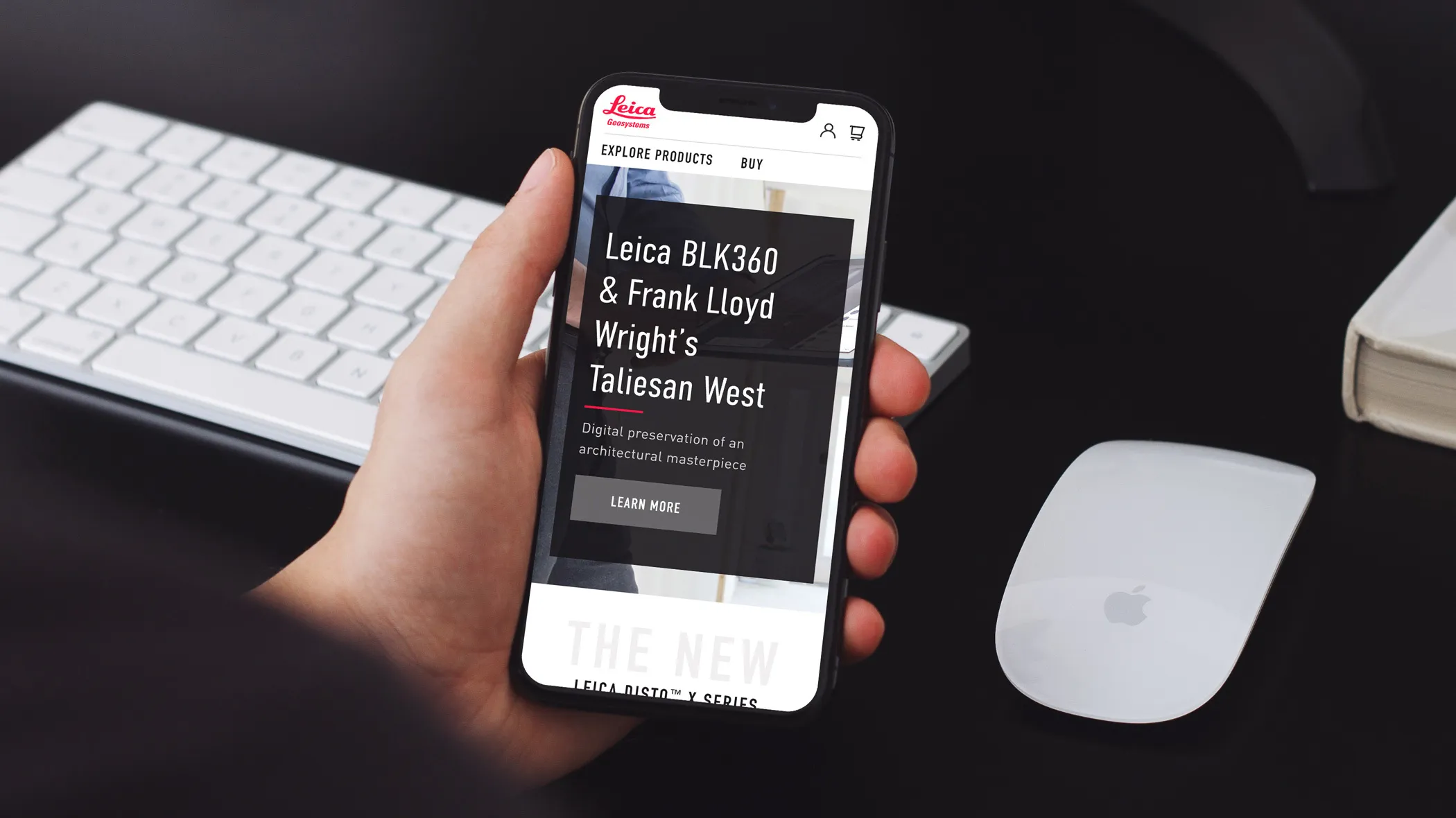 A hand at a dark desk holds a phone showing the Leica mobile site: a story titled “Leica BLK360 & Frank Lloyd Wright’s Taliesin West — digital preservation of an architectural masterpiece” above a Learn More button.