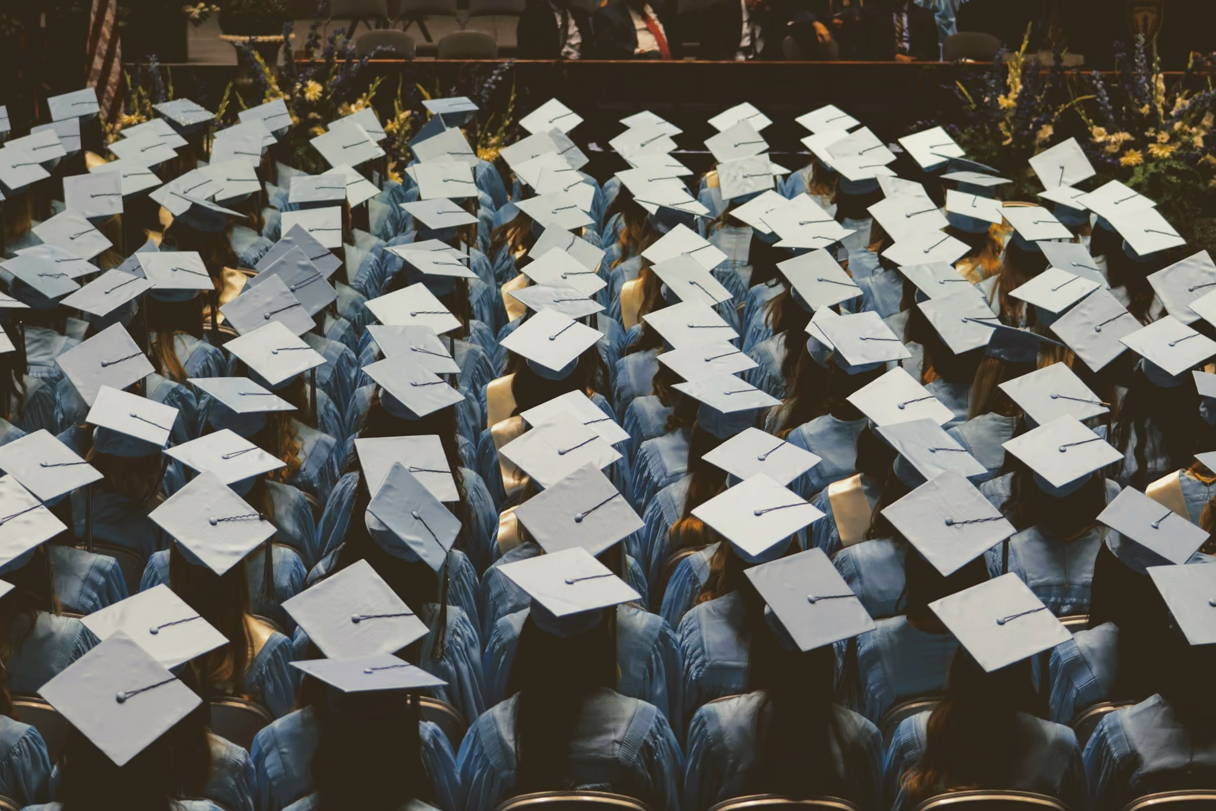 Rows of graduates in light blue gowns and pale caps, seen from behind at a high school commencement ceremony.