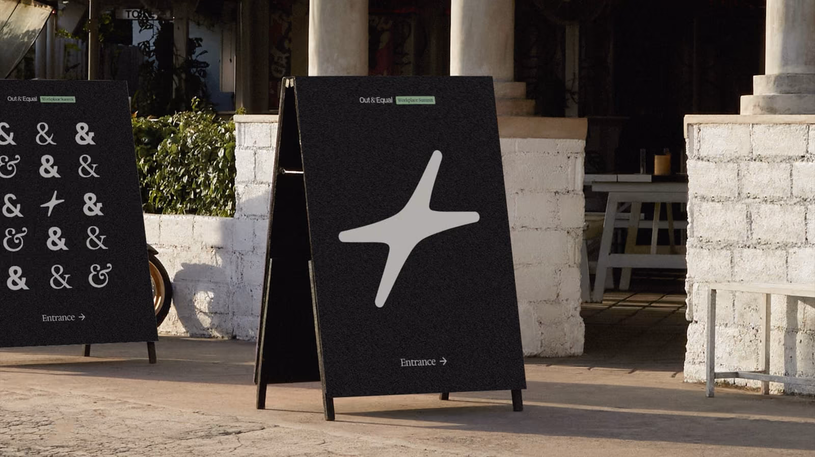 Two A-frame boards outside a summit venue in the new Out & Equal identity. One shows a grid of ampersands set in different typefaces with a four-pointed star among them; the other shows that star alone at full size, both under an “Out & Equal Workplace Summit” lockup, both reading “Entrance”.