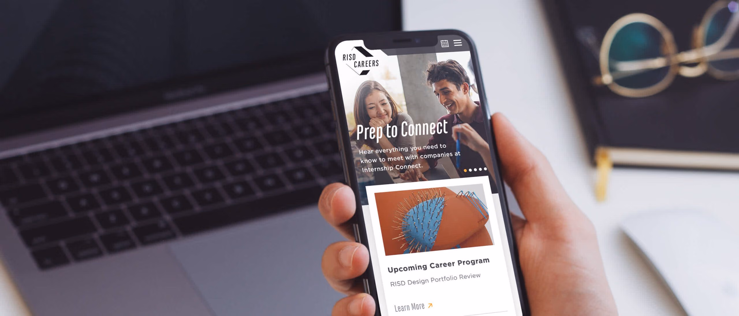 A hand holding a phone showing the RISD Careers site, a laptop and glasses out of focus behind it. The screen shows the RISD Careers mark over a photograph of two students talking, headed “Prep to Connect — Hear everything you need to know to meet with companies at Internship Connect”, above an “Upcoming Career Program” card for a RISD Design Portfolio Review.